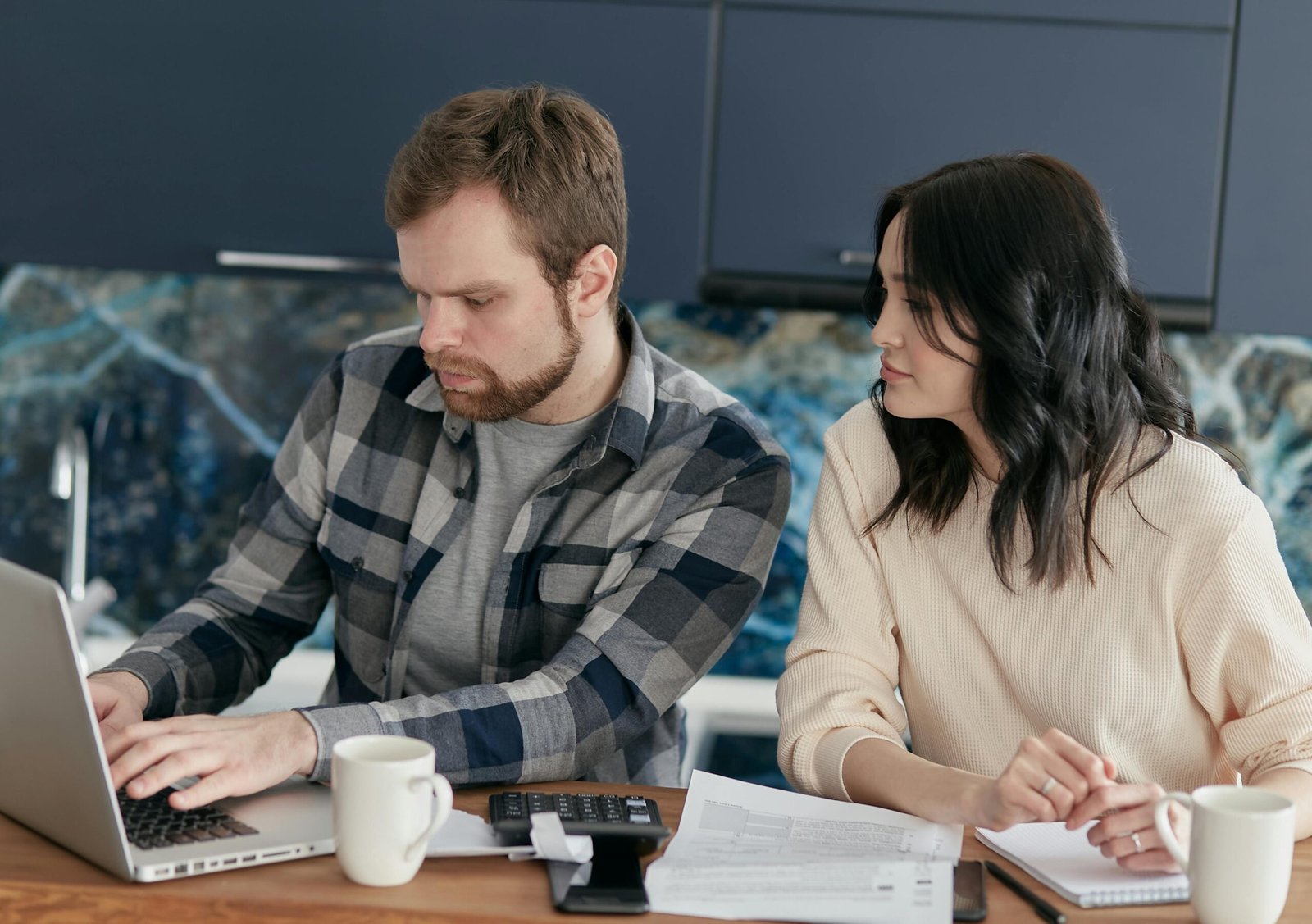 Een man en een vrouw zitten aan een houten tafel in een moderne keukenomgeving. De man typt geconcentreerd op een laptop, terwijl de vrouw over zijn schouder meekijkt naar het scherm. Voor hen liggen papieren documenten, een rekenmachine, een smartphone en twee witte koffiemokken. Ze lijken samen complexe financiële gegevens of compliance-documenten te beoordelen.