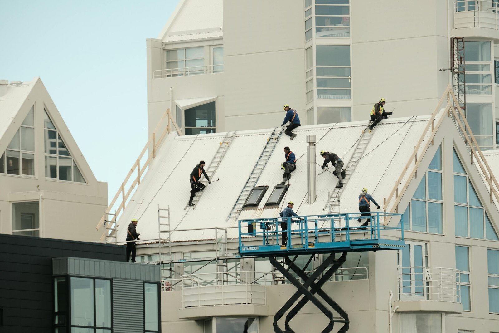 Een groep bouwmedewerkers aan het werk op een steil, wit dak van een modern appartementencomplex. De arbeiders gebruiken ladders, een hoogwerker en veiligheidslijnen. De foto illustreert de fysieke kant van de bouw- en installatiesector, waar personeelstekorten en efficiëntie-eisen momenteel een grote rol spelen.