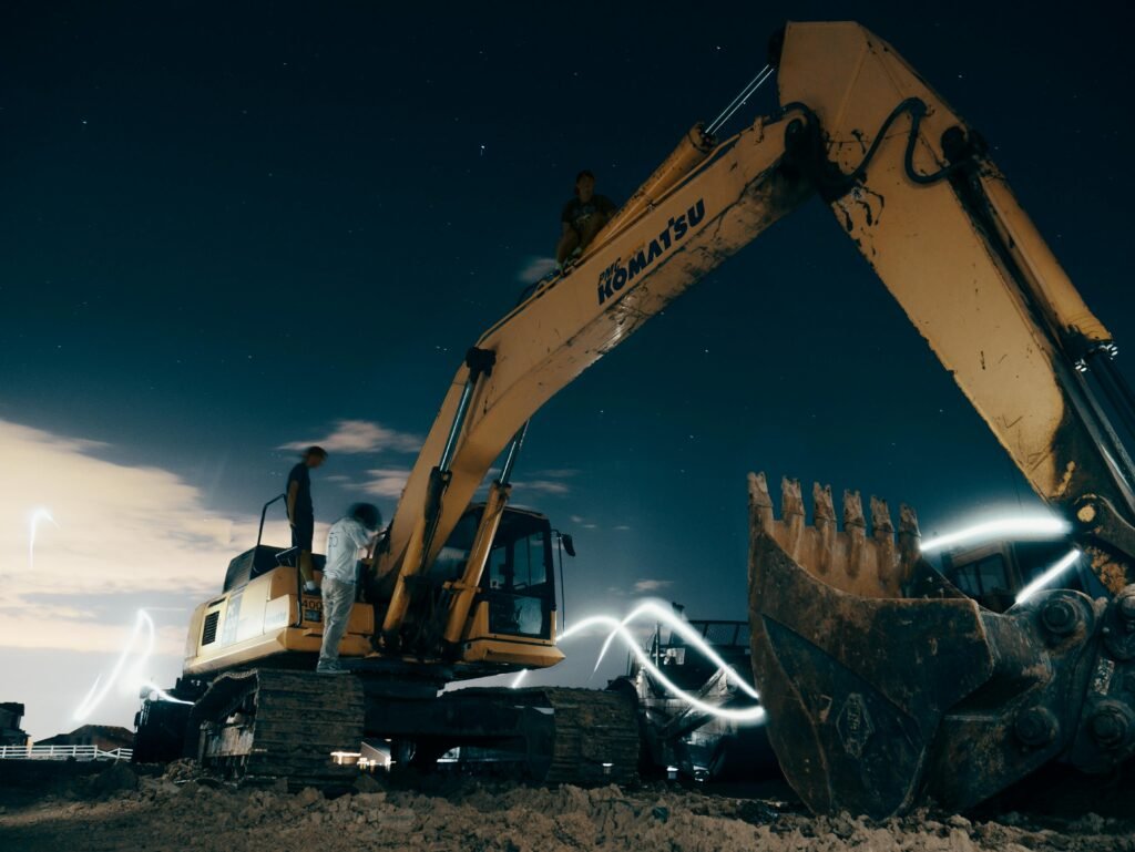 Een dramatische nachtopname van een grote gele graafmachine (Komatsu) op een bouwterrein. Lichtgevende, zwevende witte lijnen omringen de machine, wat de datastromen en algoritmes symboliseert die moderne bouwprojecten aansturen. Twee personen staan bij de machine onder een heldere sterrenhemel, wat de samenwerking tussen mens en technologie benadrukt.