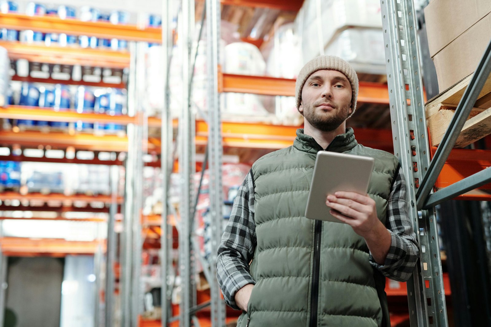 Een magazijnmedewerker met een muts en bodywarmer houdt een tablet vast terwijl hij tussen hoge stellingen met voorraad in een groot distributiecentrum staat.